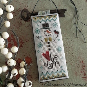 May include: A cross-stitch ornament featuring a snowman with a red heart and the text "be warm". The ornament is attached to a vintage key with a plaid ribbon.