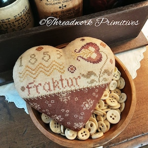 May include: A heart-shaped pillow with a brown and beige embroidered design. The pillow is decorated with a floral motif and the text "Fraktur 1853". The pillow is sitting in a wooden bowl filled with buttons.