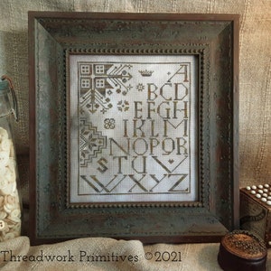 May include: A framed cross-stitch sampler with an alphabet design in brown thread on white fabric. The frame is a dark green wood with a distressed finish. The sampler is titled "Threadwork Primitives ©2021".