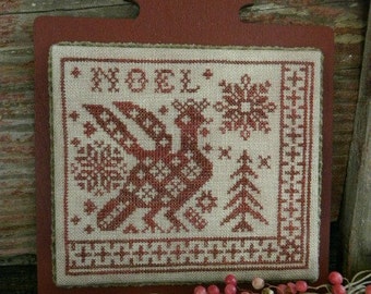 May include: A red wooden frame with a white and red cross-stitch design. The design features a bird, snowflakes, and a pine tree. The word "NOEL" is stitched in red at the top of the design.