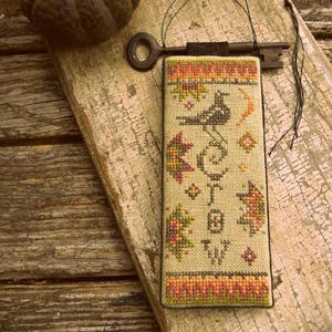 May include: A cross-stitched bookmark with a bird and floral design, a vintage key, and a small pumpkin. The bookmark has a beige background with colorful embroidery. The key is dark brown, and the pumpkin is dark green.