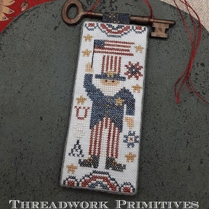 May include: A cross-stitch needlepoint ornament featuring a cartoon Uncle Sam holding a flag. The ornament is framed with a red, white, and blue border and has a keyhole at the top for hanging.