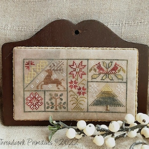 May include: A cross-stitch sampler with a winter theme. The sampler features a deer, a tree, snowflakes, and a poinsettia. The sampler is framed in a dark wood frame. The text "Threadwork Primitives © 2022" is visible at the bottom of the image.