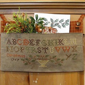 May include: A brown fabric stocking with a cross-stitch design featuring the alphabet, the word "Christmas", and the year "1747". The stocking is hanging on a wooden door with a knob.