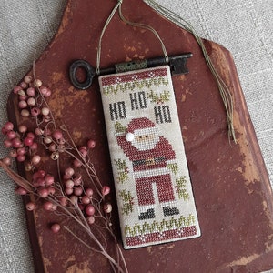 May include: A cross-stitch ornament featuring a Santa Claus figure with the text "Ho Ho Ho" on a white background. The ornament is attached to a rustic wooden board with a key.