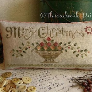 May include: A beige pillow with a pink border and the words "Merry Christmas" embroidered in brown thread. A small basket of red and orange flowers is embroidered below the text.
