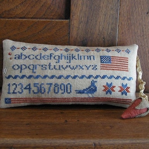 May include: A beige pillow with red, white, and blue embroidery. The pillow features the alphabet, numbers 1-900, a small bird, and a miniature American flag. The pillow is on a wooden surface.