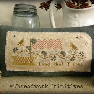May include: A cross-stitch sampler with a patriotic theme. The sampler features a red, white, and blue flag, a basket of flowers, and two birds. The text "Land that I Love" is stitched below the design.
