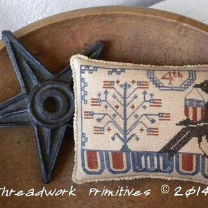 May include: A cross-stitch pillow featuring a black bird holding a small red, white, and blue bucket. The bird is perched on a branch with a tree behind it. The pillow has a blue, red, and white border with the text "4th of July" and "Threadwork Primitives © 2014".