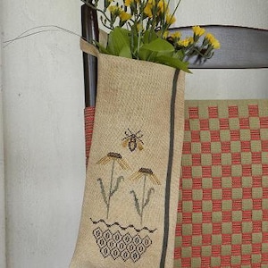 May include: A beige fabric bag with a bee and flower embroidery design. The bag is hanging on a wooden chair with a red and green checkered pattern.