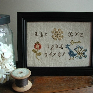 May include: A framed cross-stitch sampler with a black frame. The sampler features embroidered letters, numbers, a flower, a key, and a peacock. A glass jar filled with white buttons and a wooden spool of thread are in the foreground.