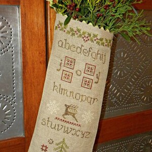 May include: A beige fabric stocking with embroidered letters, patterns, and a deer design, filled with greenery and red berries. The stocking is hanging on a wooden cabinet with metal accents. The text on the stocking reads "abcdefghij klmnopqr stuvwxyz".