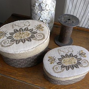 May include: Two oval-shaped, beige and brown fabric boxes with embroidered floral designs and the year "1833" on the top. The boxes are on a wooden surface.