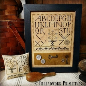 May include: A framed cross-stitch sampler with the alphabet in a vintage style font. The sampler is on a table with a small pillow featuring a similar design and the text "ABCDE F".