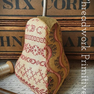 May include: A hand-stitched pear-shaped pincushion with a red and white cross-stitch design on one side and a brown and red floral fabric on the other side. The pincushion is sitting on a piece of paper with writing on it. The pincushion is in front of a wooden box with the words "SIX CORD" printed on it.