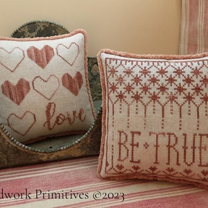 May include: Two embroidered throw pillows with pink and white fabric. The pillow on the left has a pattern of hearts and the word "love". The pillow on the right has a pattern of stars and the words "Be True".