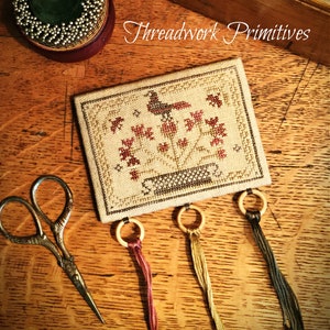 May include: A small cross-stitch sampler with a bird and flowers in a pot. The sampler is mounted on a piece of fabric and has four loops of thread hanging from it. The text "Threadwork Primitives" is visible in the background.