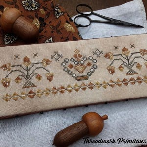 May include: A rectangular, cross-stitched piece with a beige background features acorn and floral designs in shades of brown and gold. Two wooden acorn ornaments and a pair of scissors are also present. The text "Threadwork Primitives ©2025" is visible.