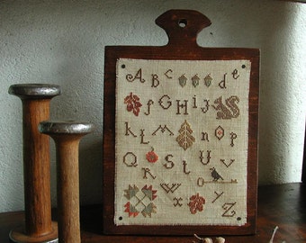 May include: A wooden framed cross-stitch sampler with the alphabet embroidered in brown and red thread. The letters are surrounded by small embroidered designs including leaves, a bird, and a pumpkin.