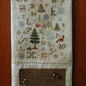 May include: A white linen needlepoint canvas with a brown felt pocket. The canvas features a cross-stitch design with a winter theme, including trees, animals, and letters. The text on the canvas reads "A C E F L M N P U V W Z".