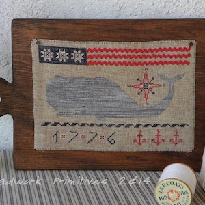 May include: A framed cross-stitch design featuring a whale with a compass and anchors. The design is on a beige fabric with red, white, and blue accents. The frame is made of dark wood.