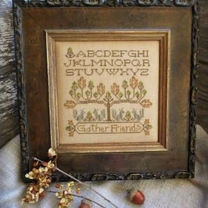 May include: A framed cross-stitch sampler with a brown wooden frame. The sampler features a tree with birds and the text "Gather Friends". The alphabet is stitched above the tree.