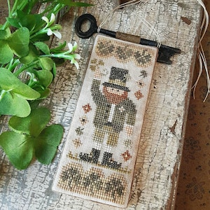 May include: A cross-stitched bookmark featuring a leprechaun design, wearing a top hat and suit, with a key attached. The bookmark has a beige background with green, brown, and orange accents. The text "Threadwork Primitives" is at the bottom.