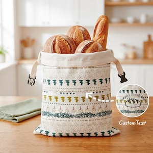 May include: A cream-colored fabric bread bag with a drawstring closure and black plastic buckles, featuring horizontal bands of green and brown patterns including trees and dots. The bag contains three loaves of bread and displays custom text.