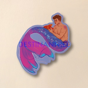 Peut inclure: Une illustration autocollante d'un triton aux cheveux bruns courts et au torse nu, tenant un petit poisson orange. Le triton a une grande queue incurvée avec un dégradé de violet, rose et bleu avec des détails scintillants. Le texte DESTINANNIE est visible.