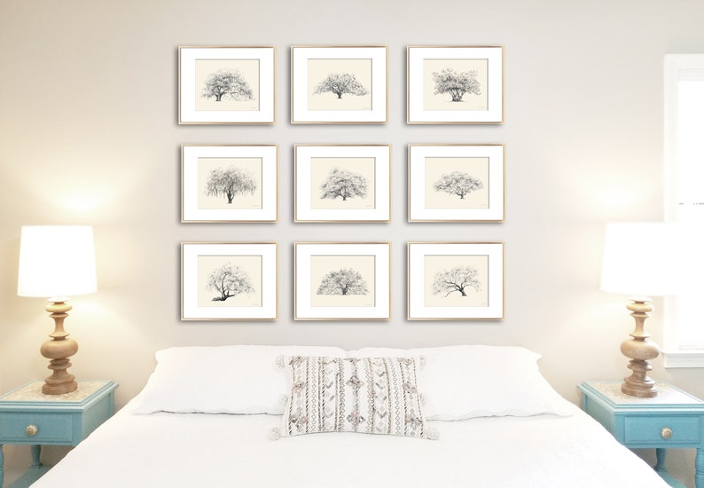 May include: A gallery wall of nine framed black and white drawings of trees, arranged in a three by three grid, above a white bed with a white pillow and a white throw pillow with a geometric pattern. Two bedside tables with lamps are on either side of the bed.