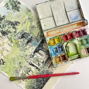 Puede incluir: Una pintura de acuarela de una fuente con &aacute;rboles y un edificio en el fondo. Una paleta de pintura de acuarela con pintura roja, amarilla, azul, verde y blanca est&aacute; abierta y un pincel rojo est&aacute; encima de la pintura.