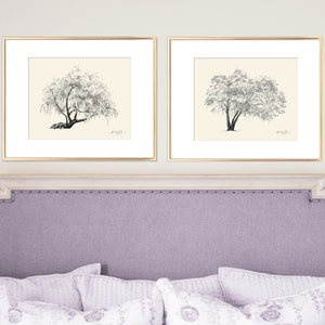 May include: Two black and white drawings of trees in gold frames are hung above a light purple headboard with a tufted design.