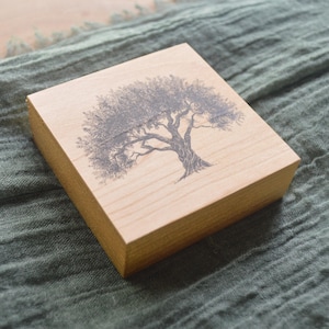 May include: A wooden block with a black and white illustration of a tree on it. The block is a light brown color and has a smooth surface.