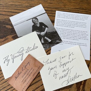 May include: A business card with the text "Fly Young Studio" and "Heather L. Young" in cursive script. The card is made of wood and has a light brown color. The card also includes a website address, phone number, and email address.