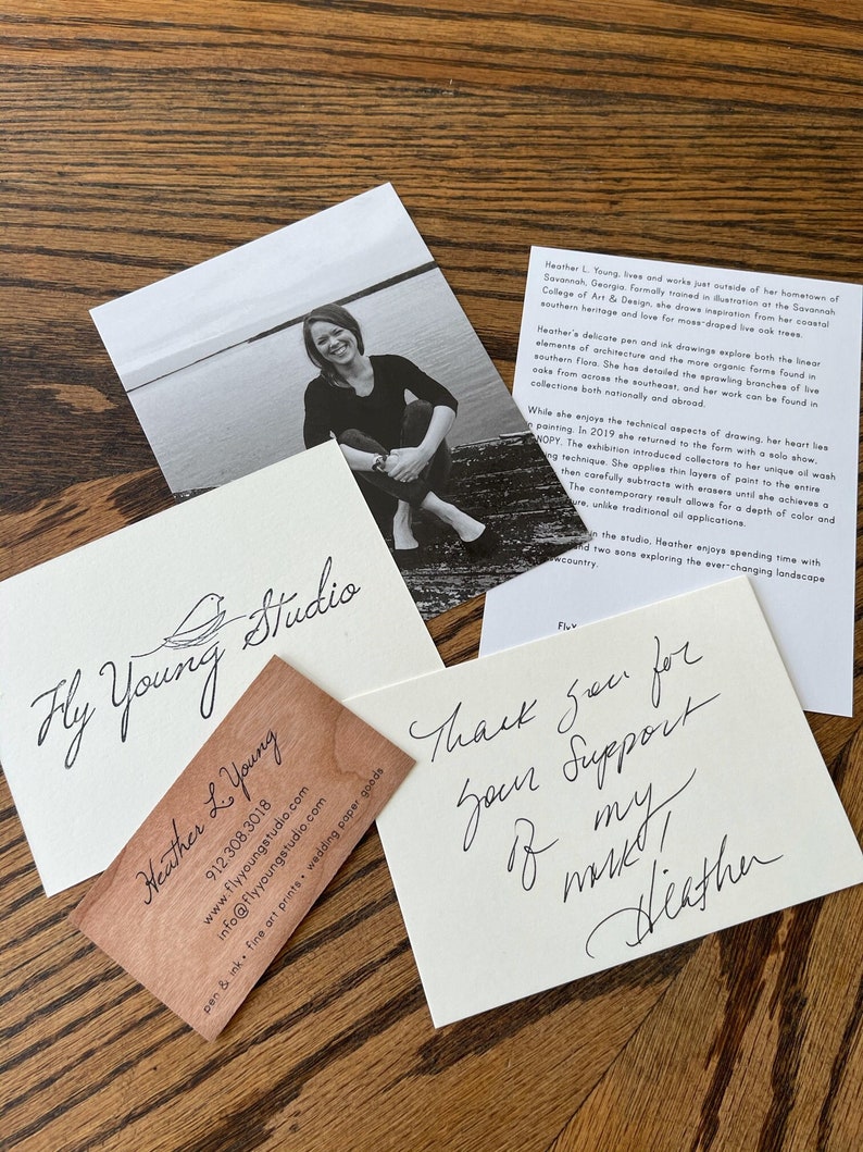 May include: A business card with the text "Fly Young Studio" and "Heather L. Young" in cursive script. The card is made of wood and has a light brown color. The card also includes a website address, phone number, and email address.