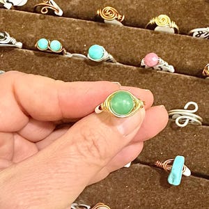 May include: A collection of handmade wire-wrapped rings with various gemstone beads. The rings feature silver and gold-colored wire, with stones in shades of green, turquoise, pink, and white. The rings are displayed on a brown surface.