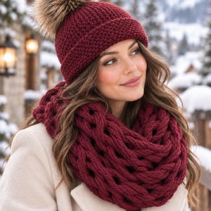 Peut inclure: Un bonnet en tricot bordeaux profond doté d'un grand pompon en fausse fourrure duveteux sur le dessus, associé à une écharpe infinie assortie en tricot épais à mailles larges, portée sur un manteau beige clair.