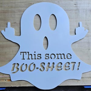 Peut inclure: Une découpe de fantôme en métal bleu clair avec le texte "This some BOO-SHEET!". Le fantôme fait un doigt d'honneur avec les deux mains. La découpe est posée sur une surface en bois.