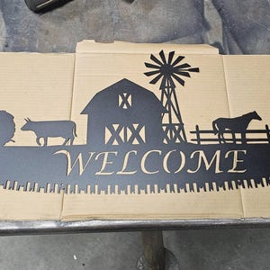May include: Black metal farm-themed sign with a saw blade design. The sign features a tractor, cow, barn, windmill, horse, and corn stalks. The word "WELCOME" is cut out in the centre of the sign.