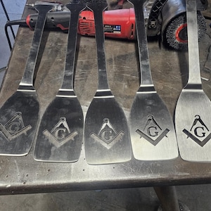 Könnte beinhalten: Fünf Metallspatel mit einem Freimaurersymbol auf der Klinge. Die Spatel haben lange, schmale Griffe und breite, abgerundete Klingen. Die Klingen zeigen das Symbol von Zirkel und Winkel mit dem Buchstaben "G" in der Mitte. Die Spatel sind aus glänzendem, silberfarbenem Metall.