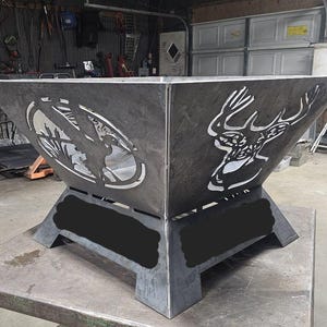 以下が含まれることがあります： 濃いグレー仕上げの、大きな正方形の金属製ファイヤーピット。側面には、鹿と狩人の切り絵のデザインが施されています。ファイヤーピットは頑丈なベースを備え、屋外での使用を目的としています。 全体的なデザインは素朴です。