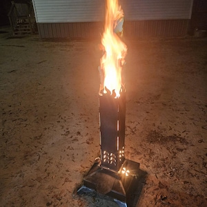 Puede incluir: Un brasero alto de metal negro con llamas que se elevan desde la parte superior. El brasero tiene cortes decorativos y una base cuadrada. Las llamas son de color naranja brillante y amarillo contra el cielo nocturno. El brasero está al aire libre.