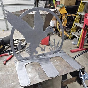 May include: A metal sculpture of a duck in flight, set within a circular frame. The sculpture features cattails and a base with two curved supports. The piece is made of raw metal and appears to be hand-welded.