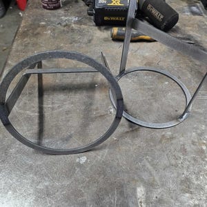 Pode incluir: Trabalho em metal em andamento: Duas estruturas circulares de metal com estruturas de suporte, possivelmente para um item decorativo ou funcional. O metal inacabado tem uma aparência bruta e industrial, sugerindo um design feito à mão ou personalizado. As peças estão em uma superfície de metal.