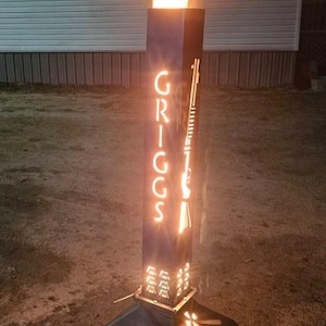 May include: A tall, black metal fire pit with a burning flame. The fire pit has the name "GRIGGS" cut out on one side and a rifle silhouette on the other. The base is a dark, geometric shape.