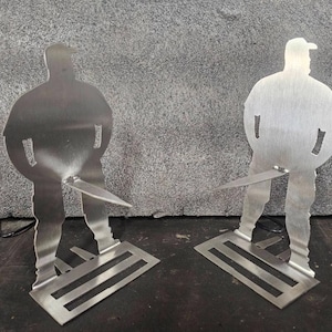 Könnte beinhalten: Zwei silberne Metall-Silhouette-Figuren einer Person mit Hut, Händen in den Taschen und einem Messer. Die Figuren stehen auf einer rechteckigen Basis mit drei horizontalen Linien. Der Hintergrund ist eine graue Wand.