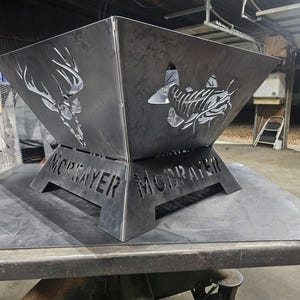 May include: A custom metal fire pit with cut-out designs of a deer and a fish. The fire pit is dark grey and has the name "MCBRAYER" cut into the base. It is sitting on a metal surface.