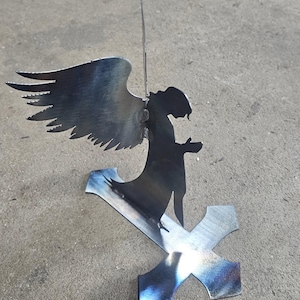 Puede incluir: Una escultura de metal de un ángel con las alas extendidas, de pie sobre una cruz. El ángel está silueteado contra un fondo claro, con una espada que se extiende hacia arriba. El metal tiene un aspecto oscuro y texturizado.