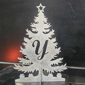 May include: A silver-colored metal Christmas tree decoration with a star on top and the letter "Y" in the center. The tree has a cutout design and stands upright with a base for support.