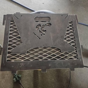 May include: A dark gray metal fire pit grate with a cut-out duck design. The grate features a diamond-patterned mesh and a handle. The overall design is square with tabs on the sides.